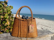 A brown woven handbag with a tag attached, placed on a wooden surface near a beach with a clear blue sky in the background.