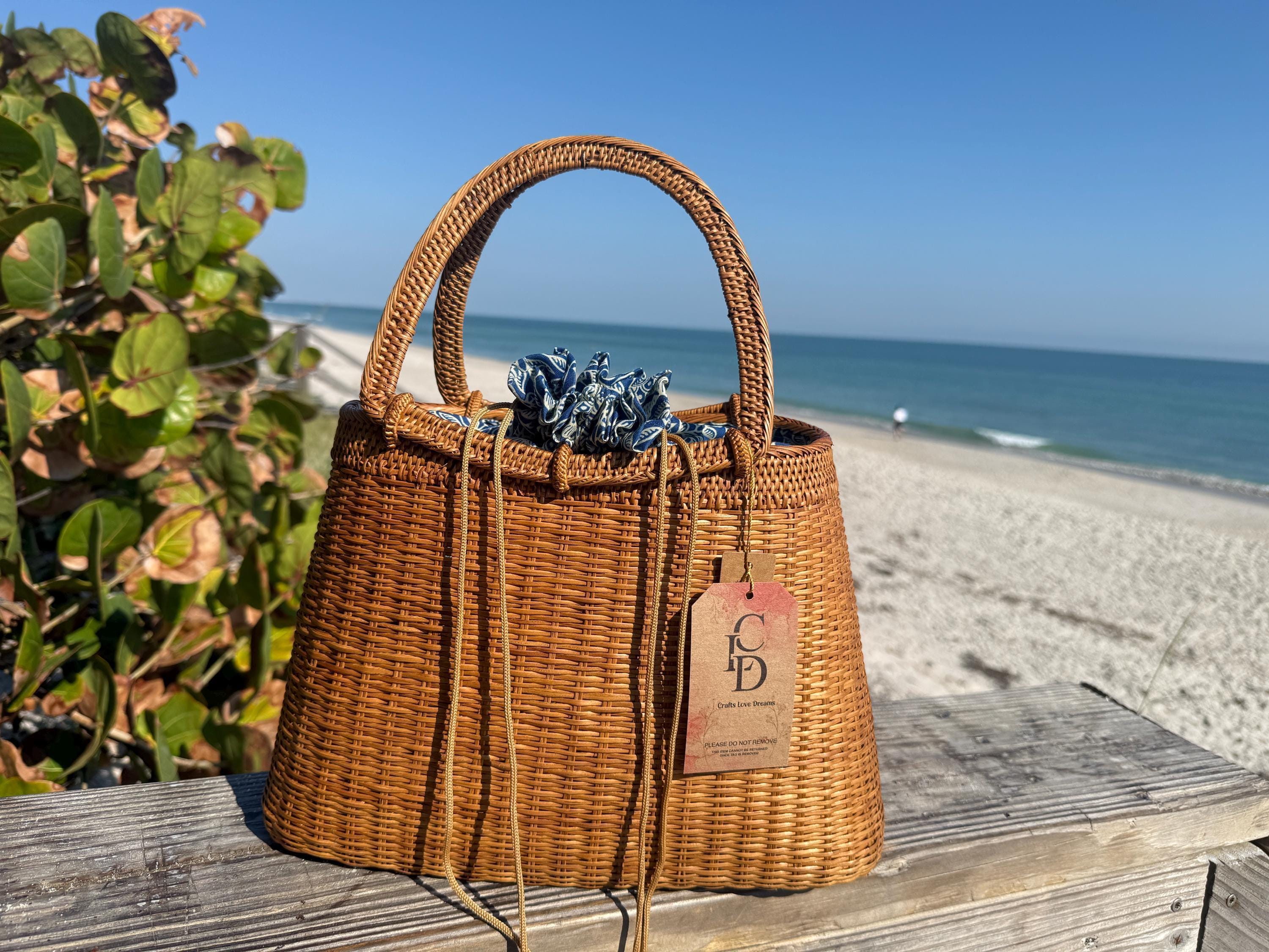 A brown woven handbag with a tag attached, placed on a wooden surface near a beach with a clear blue sky in the background.