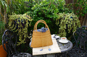 A woven straw handbag sits on a metal table, surrounded by potted plants and a white teacup, creating a cozy and inviting outdoor setting.