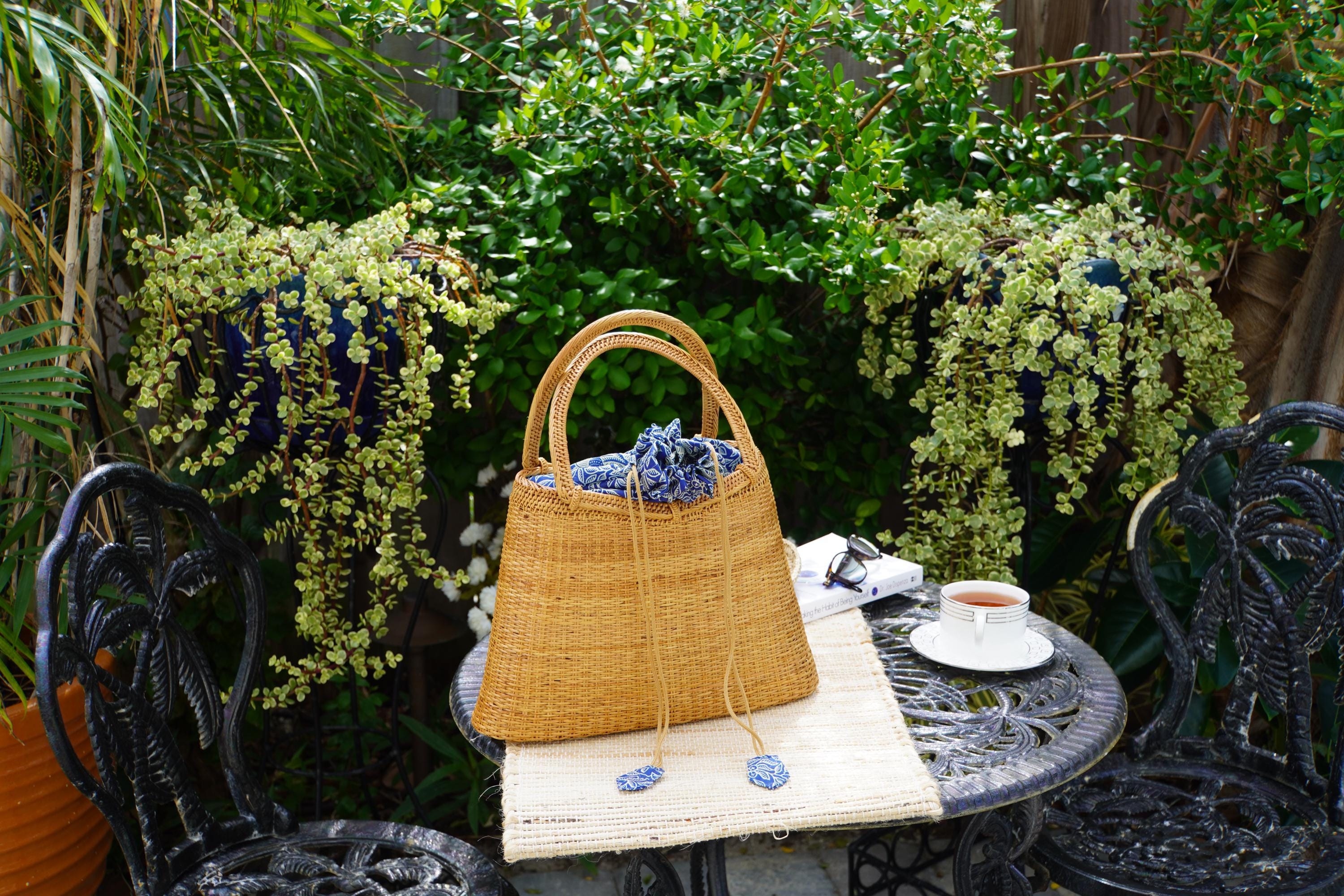 A woven straw handbag sits on a metal table, surrounded by potted plants and a white teacup, creating a cozy and inviting outdoor setting.