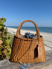 A brown woven handbag with a blue and white floral pattern sits on a wooden surface, with a beach and clear blue sky in the background.
