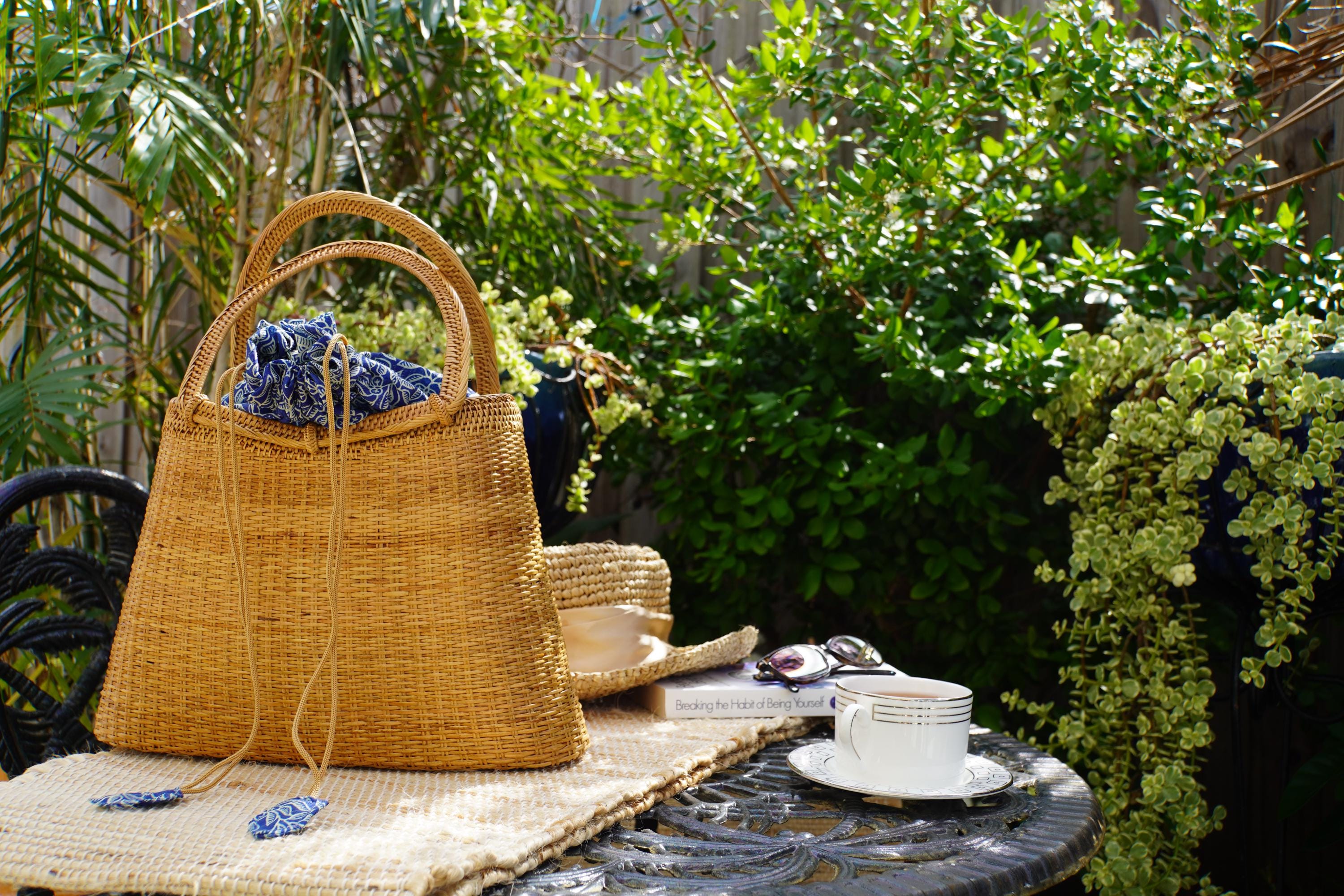 A woven straw bag with blue accents sits on a woven mat, accompanied by a white teacup, a book, and a hat on a table in a lush garden setting.