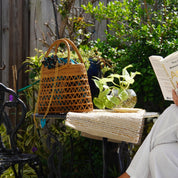 A person is sitting on a black wrought iron chair, reading a book, with a woven purse and a potted plant nearby.