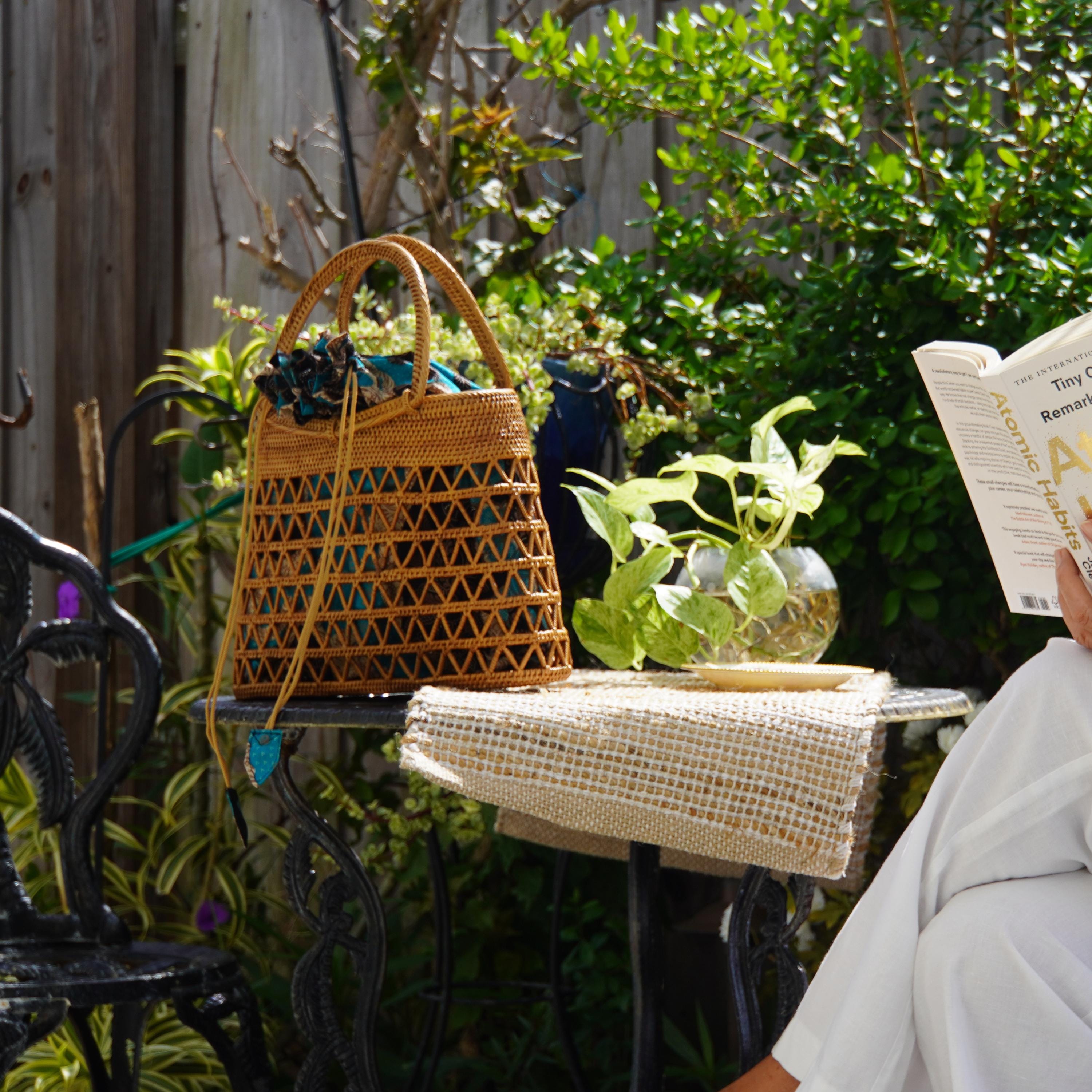 A person is sitting on a black wrought iron chair, reading a book, with a woven purse and a potted plant nearby.