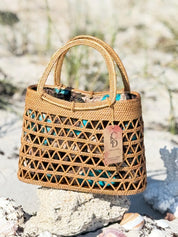 A woven handbag with a distinctive pattern sits on a rock in a sandy beach setting.