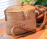 A woven straw handbag with a leather strap, displayed on a wooden surface.