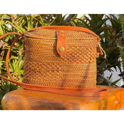 A woven straw bag with a leather strap, placed on a wooden surface against a backdrop of lush greenery.