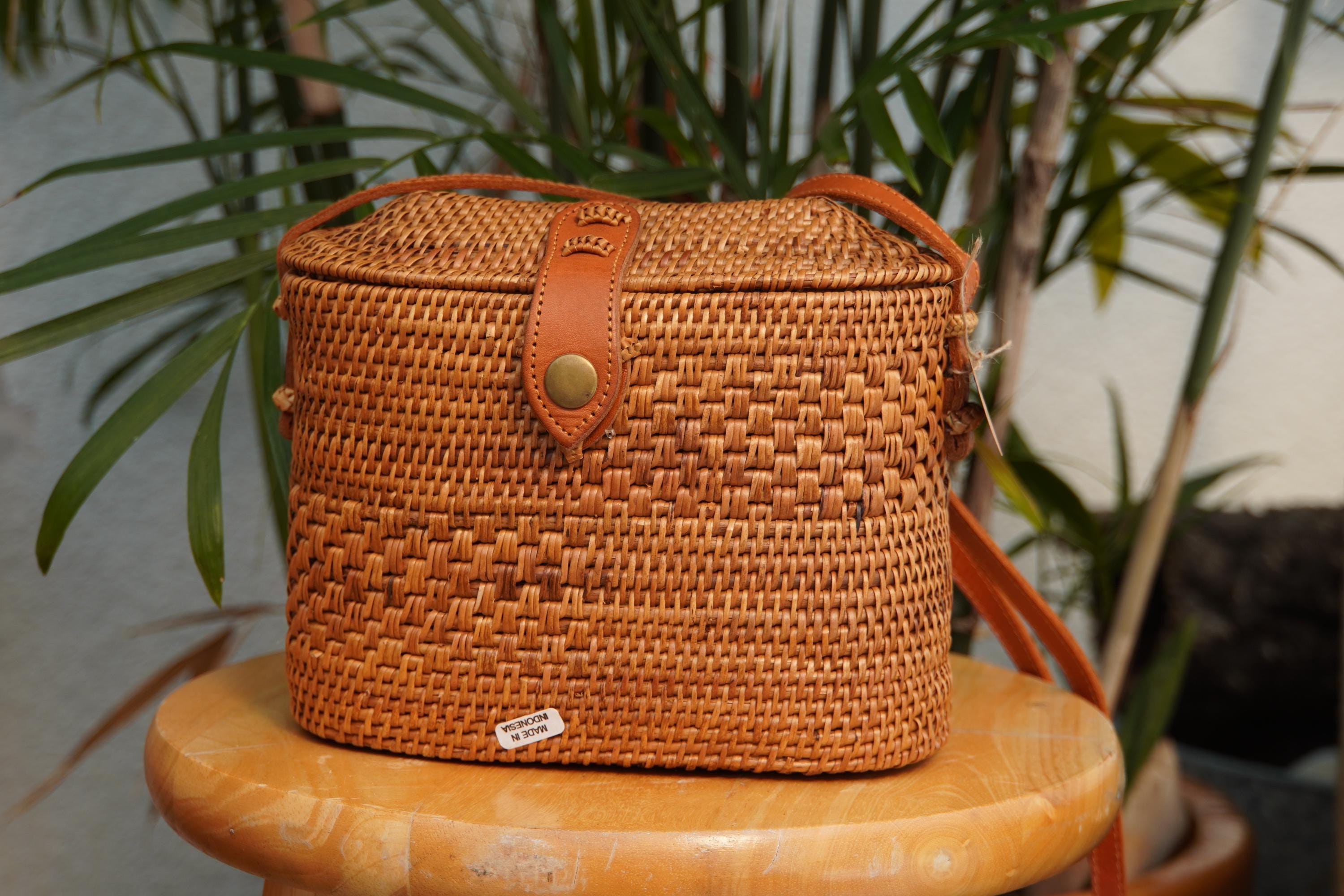 A brown woven handbag with a leather strap is displayed on a wooden surface, with a plant in the background.