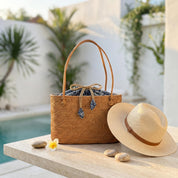 A woven straw bag with a blue pattern sits on a table next to a straw hat, with a small flower and some rocks nearby.