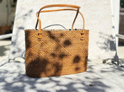 A woven straw handbag with a brown leather handle and strap is placed on a white chair, casting a shadow on the chair's surface.
