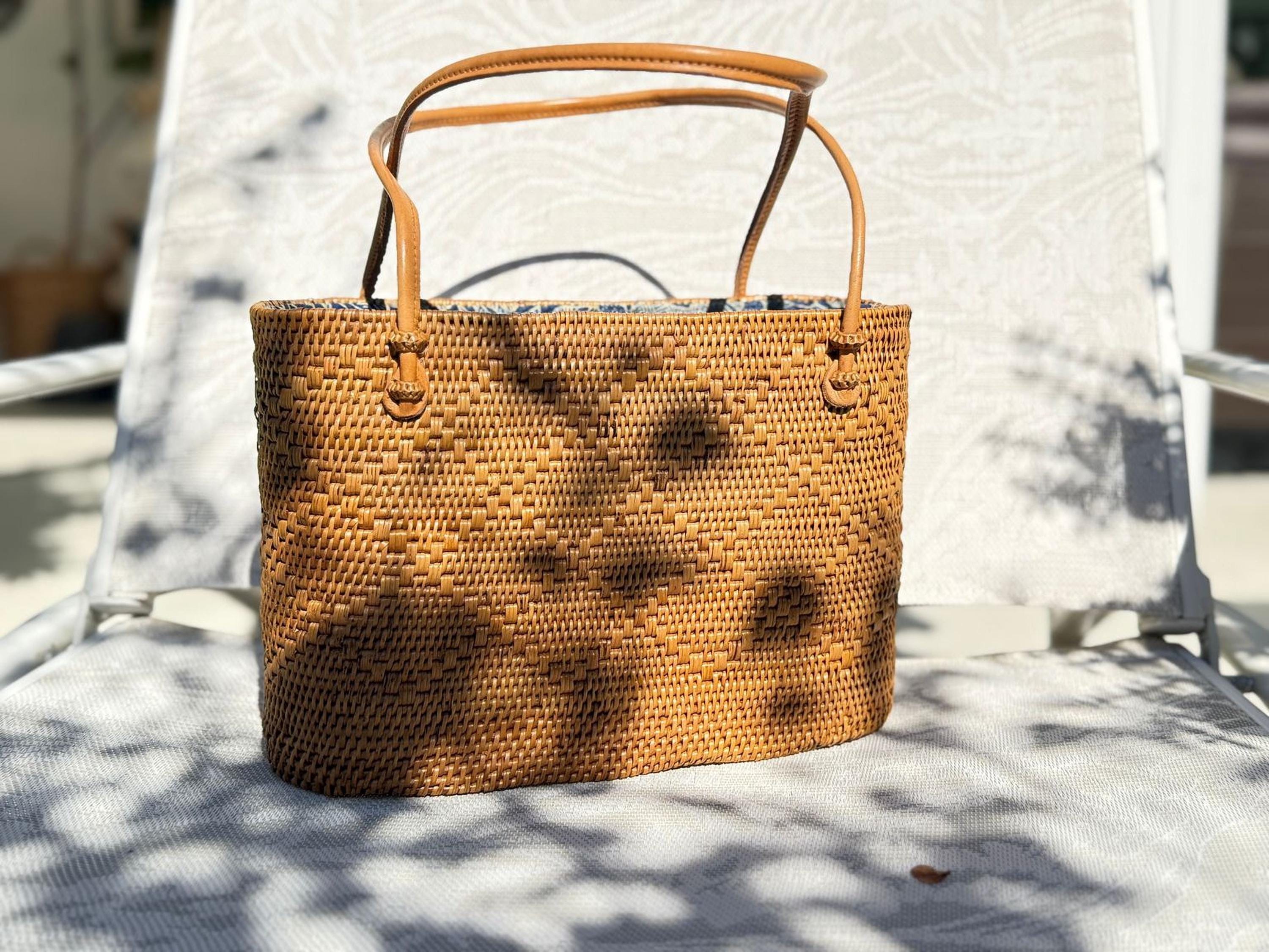 A woven straw handbag with a brown leather handle and strap is placed on a white chair, casting a shadow on the chair's surface.