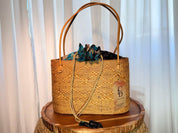 A woven straw handbag with a blue and black patterned fabric, placed on a wooden surface against a white curtain background.
