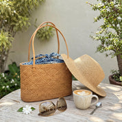 A woven straw bag with a blue and white pattern sits on a wooden table, accompanied by a straw hat, sunglasses, and a cup of coffee.