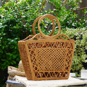A woven straw bag with a round handle and a patterned design sits on a table in a garden setting.