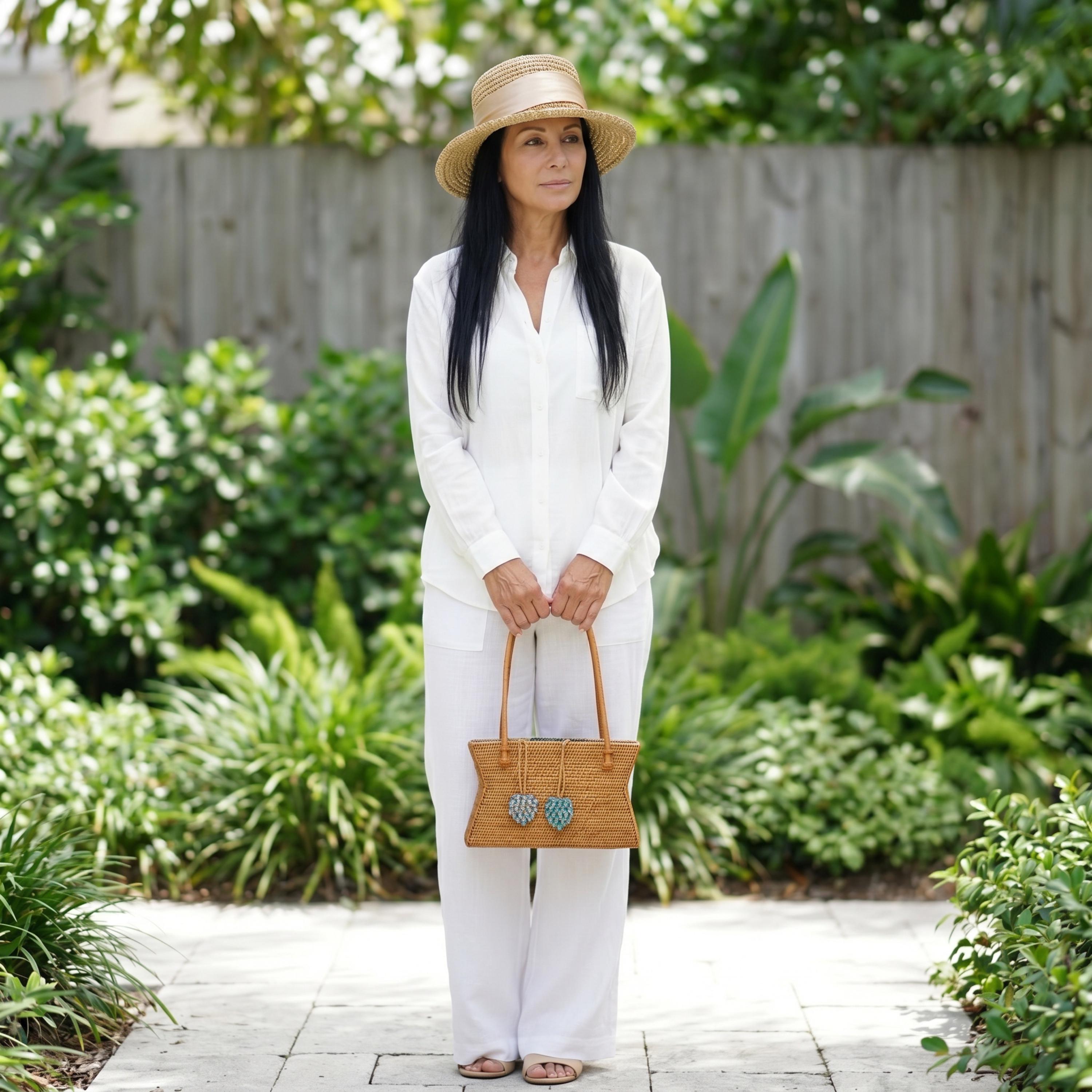 A woman in a white outfit and straw hat stands in a garden, holding a brown purse with heart-shaped charms.