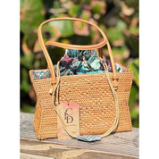 A woven straw handbag with a patterned fabric lining sits on a wooden surface, with a tag attached to it.