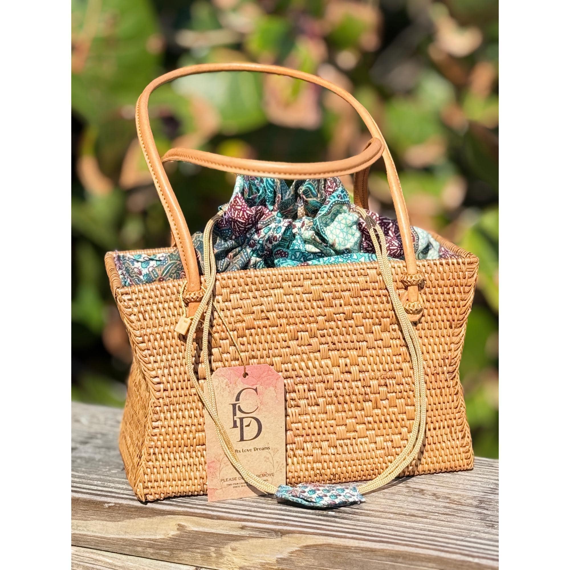 A woven straw handbag with a patterned fabric lining sits on a wooden surface, with a tag attached to it.