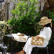 A woman in a white outfit and straw hat sits on a bench, holding a star-patterned purse, with a small table and plants in the background.