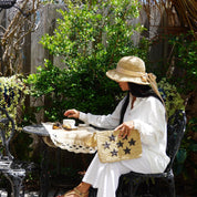 A woman in a white outfit and straw hat sits at a table with a woven bag, enjoying a drink in a garden setting with lush greenery and a wooden fence in the background.