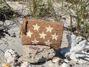 A woven purse with star-shaped embellishments rests on a rock in a sandy beach setting, surrounded by seashells and a piece of coral.