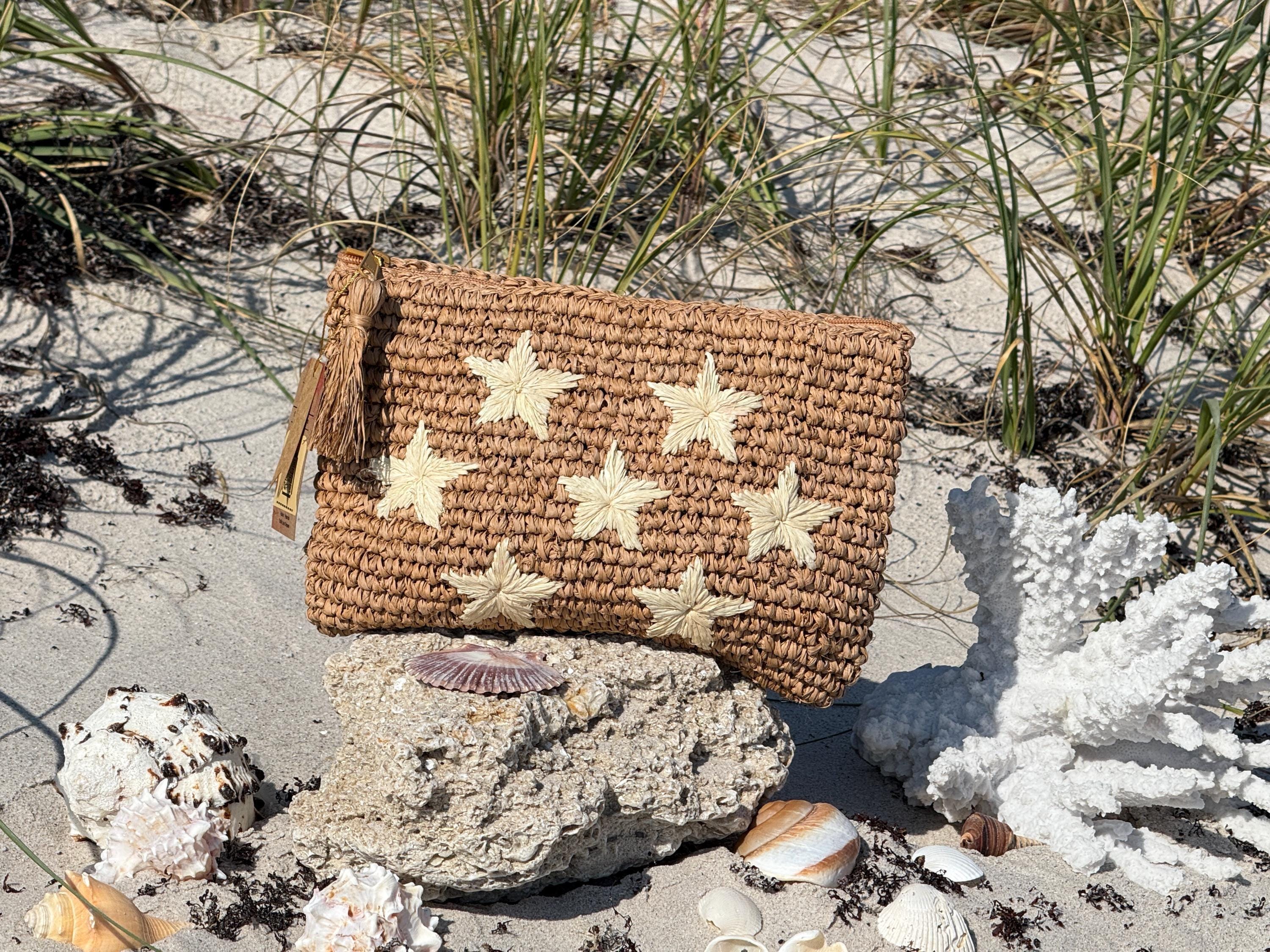 A woven purse with star-shaped embellishments rests on a rock in a sandy beach setting, surrounded by seashells and a piece of coral.