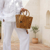 A person wearing white pants and holding a woven handbag stands in front of a stone wall with a potted plant.