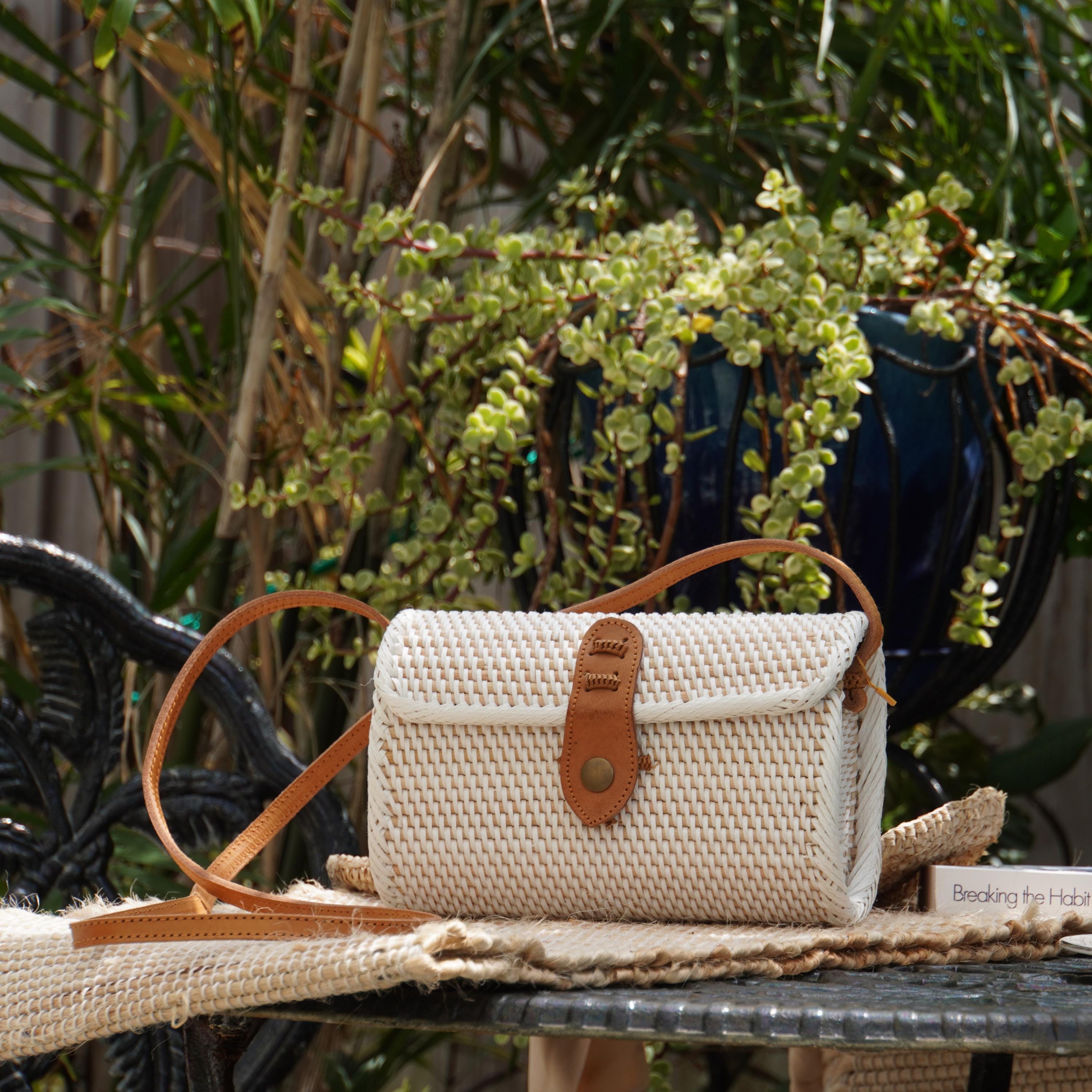 A white woven purse with a leather strap is placed on a table, with a potted plant and a black chair visible in the background.