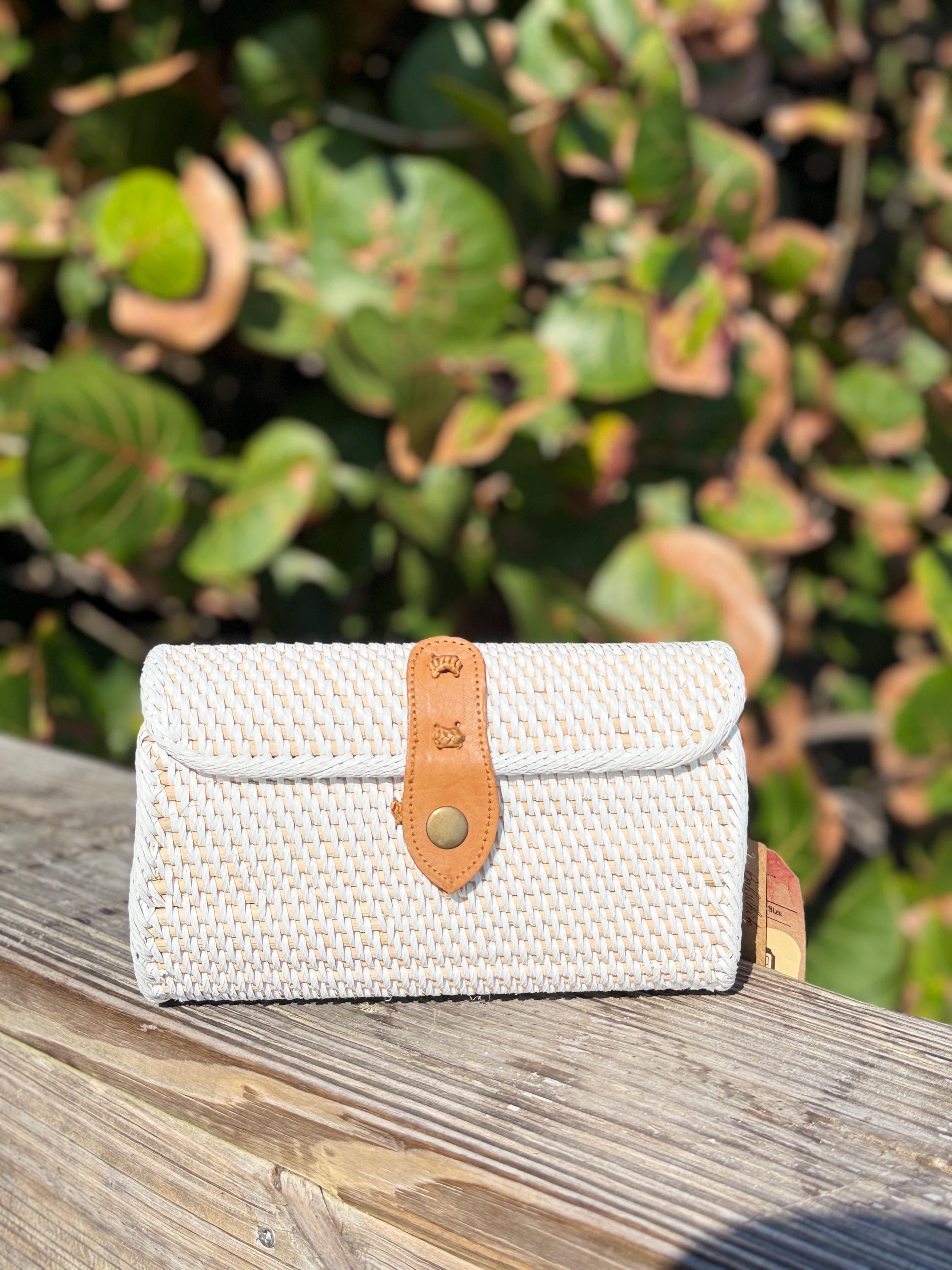 A white woven purse with a leather strap is placed on a wooden surface, with a blurred background of green leaves.