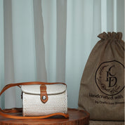A white woven purse with a leather strap and a burlap sack with a logo are displayed on a wooden surface, with a white curtain in the background.