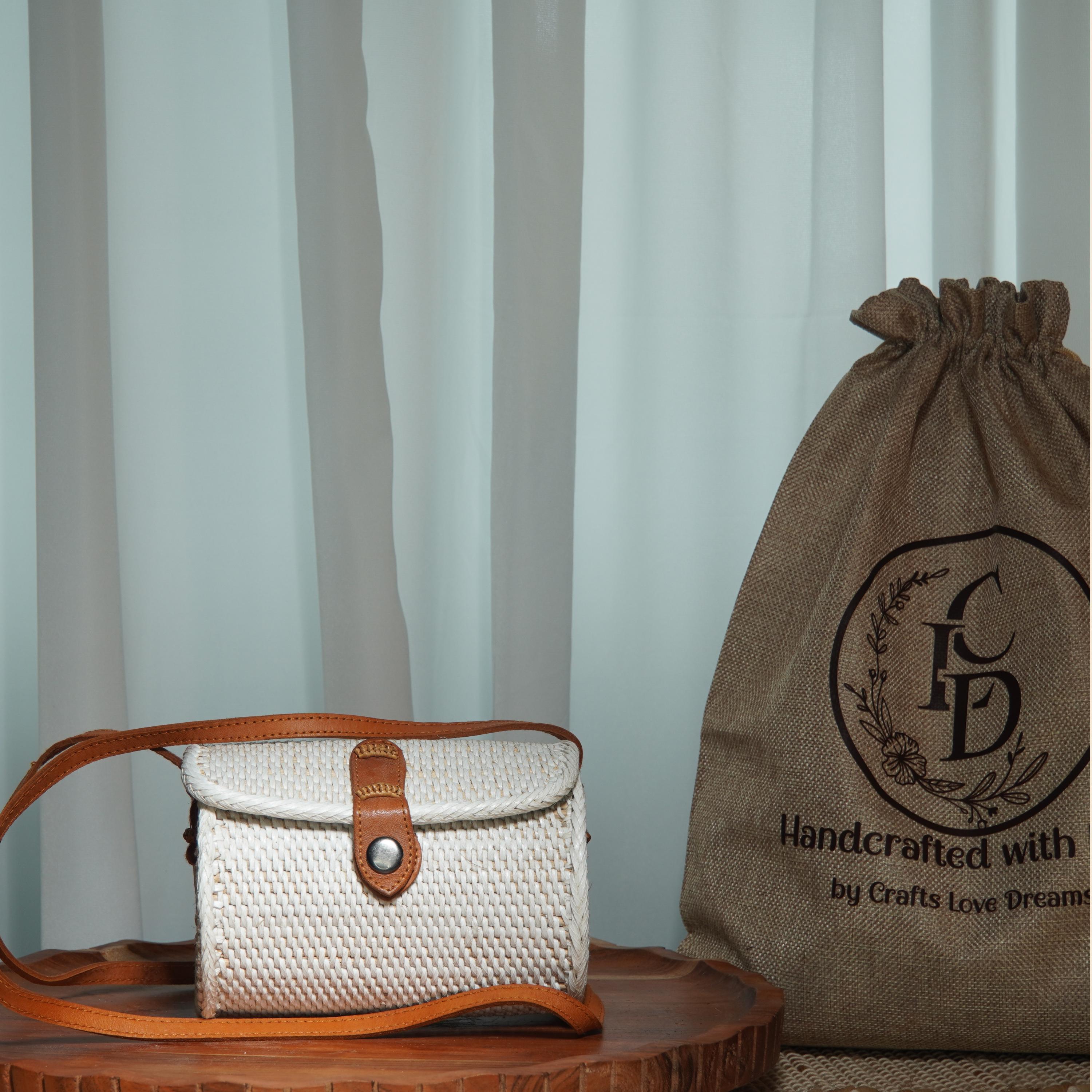 A white woven purse with a leather strap and a burlap sack with a logo are displayed on a wooden surface, with a white curtain in the background.