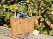 A woven straw handbag with a floral pattern sits on a wooden surface, accompanied by a seashell and a necklace.