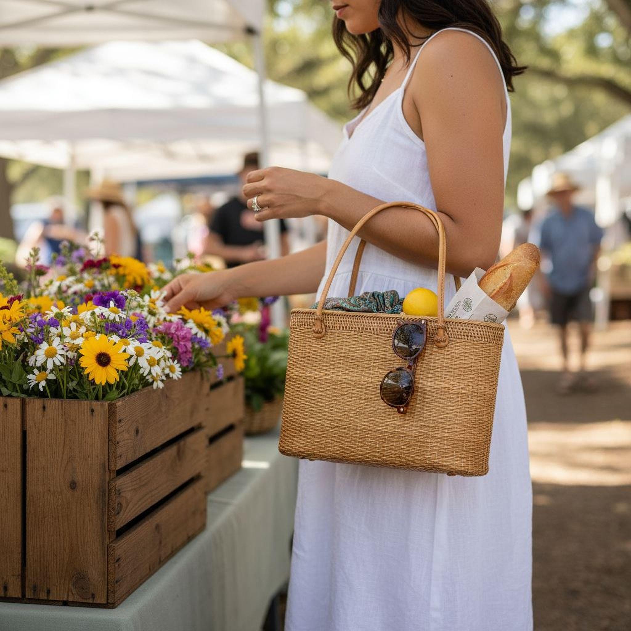 A woman in a white dress is browsing through a basket of flowers at an outdoor market, with a straw bag containing sunglasses and a loaf of bread in her hand.
