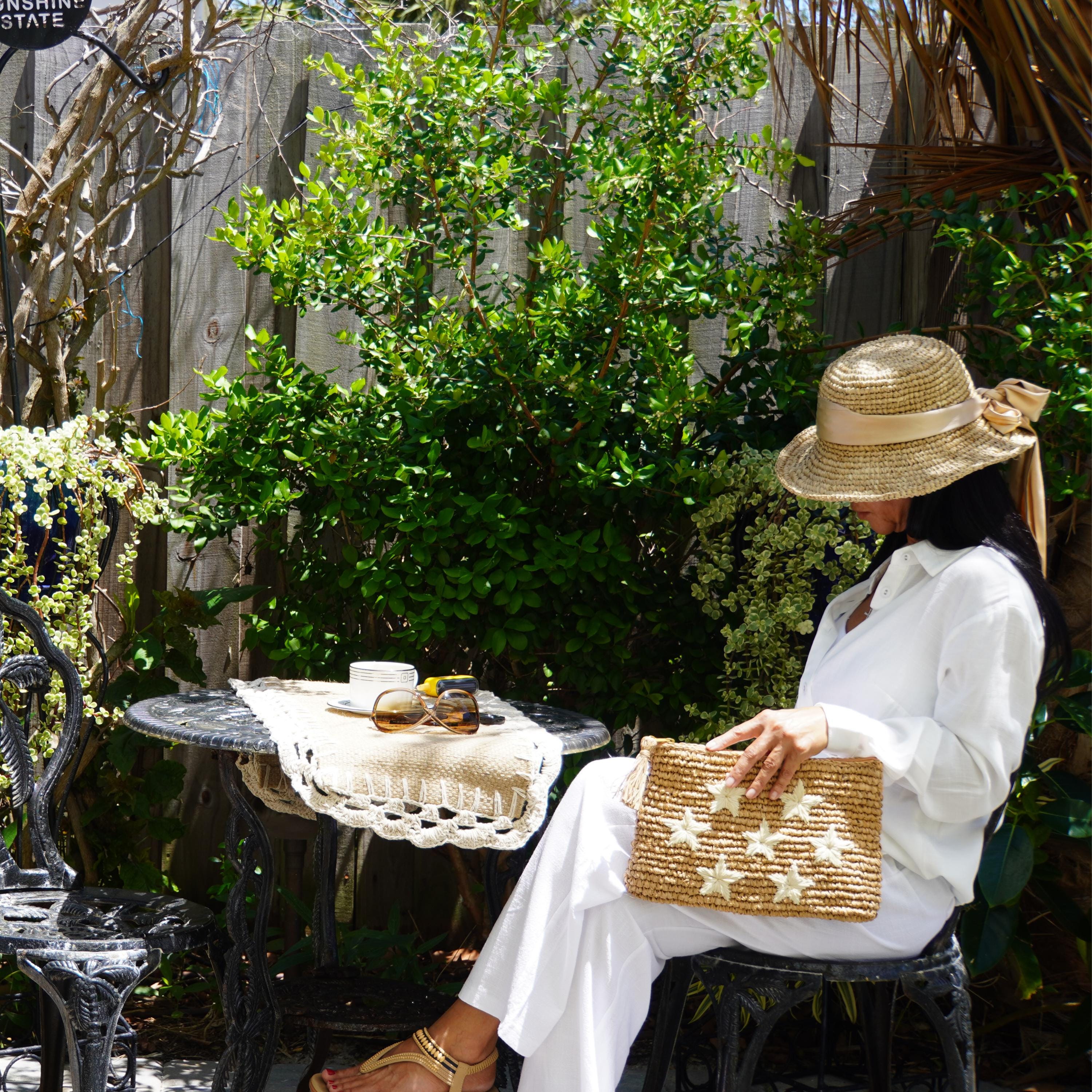 A woman in a white outfit and straw hat sits on a bench in a lush garden, holding a star-patterned purse.