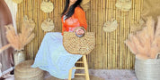 A woman in a red top and white skirt is sitting on a wooden stool in front of a bamboo wall with decorative leaves.
