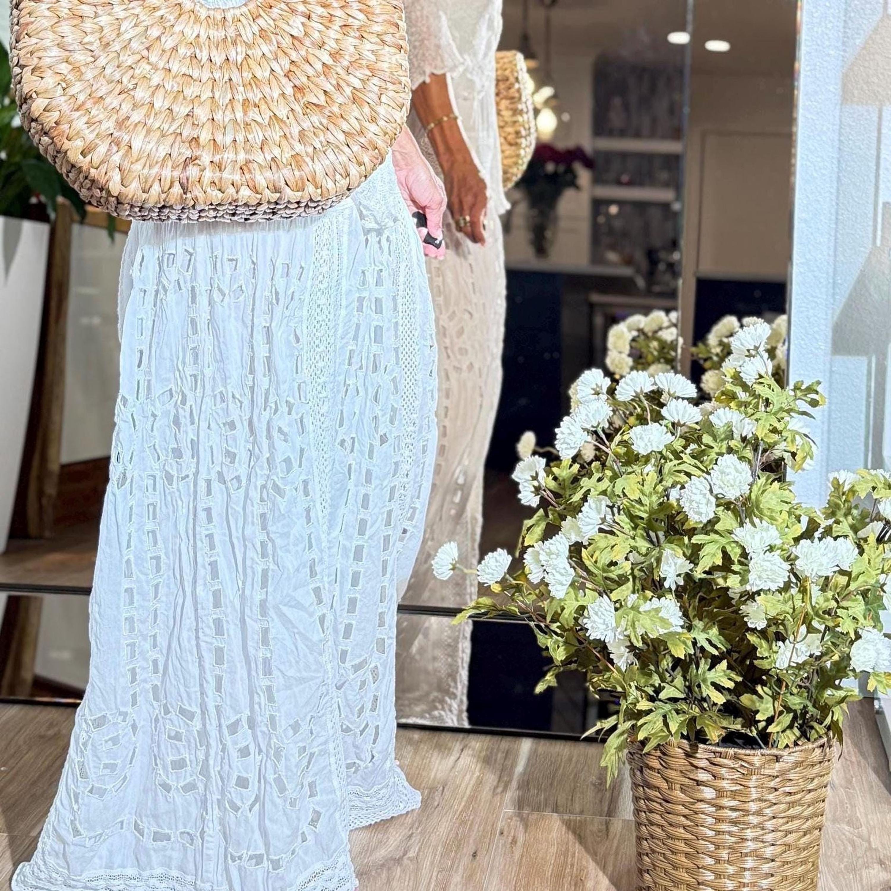 a woman wearing a white lace dress standing next to a potted plant with white flowers. The potted plant is placed on a wooden floor, and there is a mirror in the background.