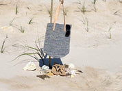 A blue and white striped tote bag with sunglasses hanging from it, placed on a sandy beach with some sandals and seashells nearby.